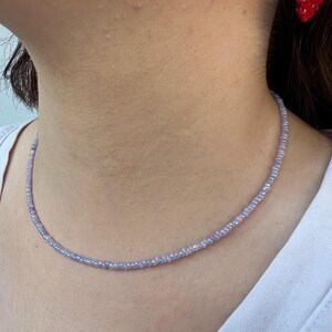K&ouml;nnte beinhalten: Eine zarte, hellviolette Perlenkette. Die Halskette besteht aus kleinen, gleichm&auml;&szlig;ig verteilten Perlen, die einen dezenten, eleganten Look erzeugen. Ein roter und gr&uuml;ner Erdbeerohrring ist im Hintergrund zu sehen.