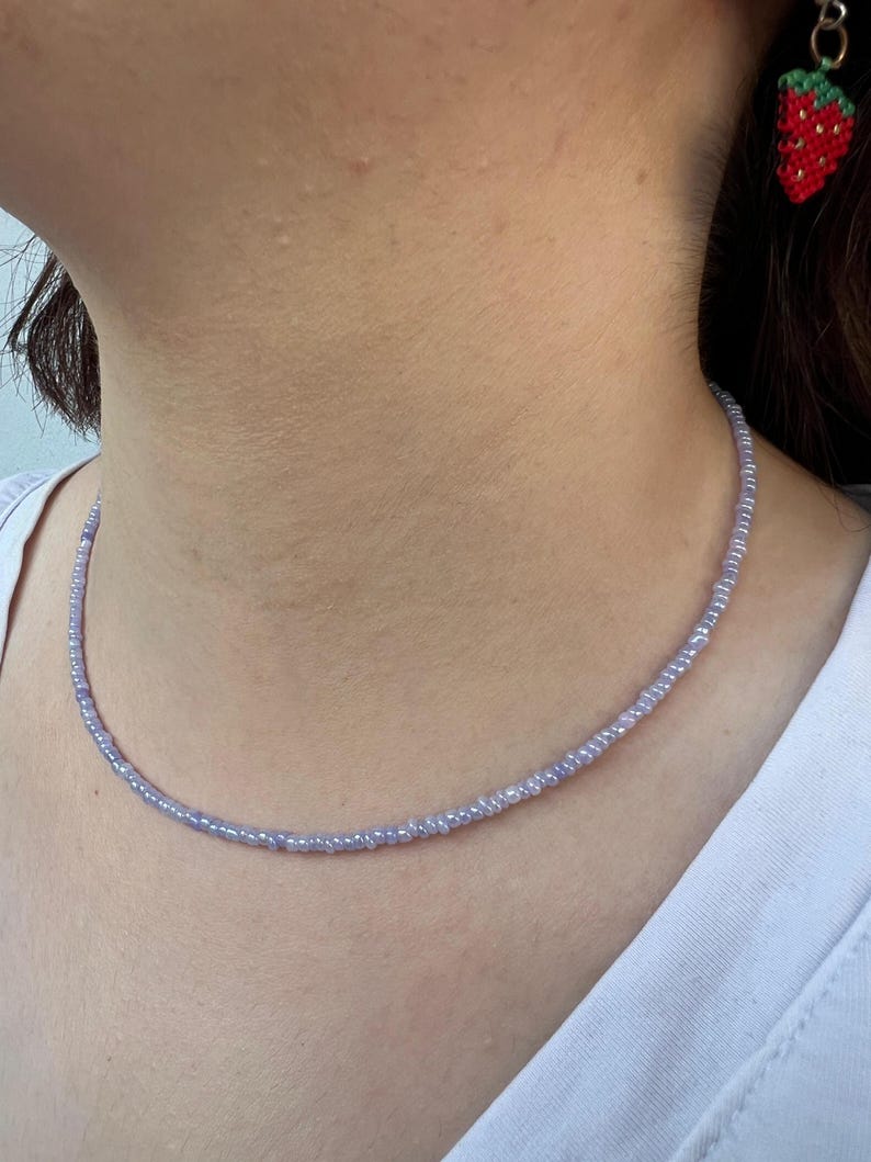 K&ouml;nnte beinhalten: Eine zarte, hellviolette Perlenkette. Die Halskette besteht aus kleinen, gleichm&auml;&szlig;ig verteilten Perlen, die einen dezenten, eleganten Look erzeugen. Ein roter und gr&uuml;ner Erdbeerohrring ist im Hintergrund zu sehen.
