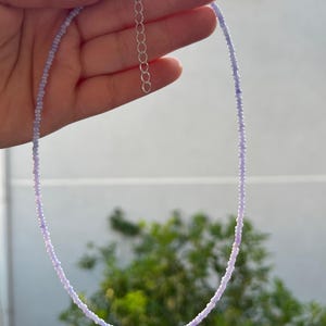 K&ouml;nnte beinhalten: Eine zarte Halskette mit kleinen, hellvioletten Perlen, die aufgef&auml;delt sind. Die Halskette hat einen silberfarbenen Verschluss und eine Verl&auml;ngerungskette. Die Perlen gehen von einem dunkleren zu einem helleren Violett &uuml;ber. Die Kette wird vor einem verschwommenen gr&uuml;nen Hintergrund gehalten.