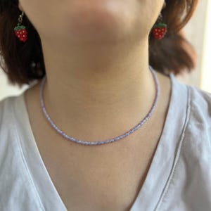 K&ouml;nnte beinhalten: Eine zarte, hellviolette Perlenkette wird getragen. Die Kette wird mit Erdbeerohrringen kombiniert. Die Ohrringe sind rot mit gr&uuml;nen Bl&auml;ttern und wei&szlig;en Punkten. Die Person tr&auml;gt ein wei&szlig;es Hemd.