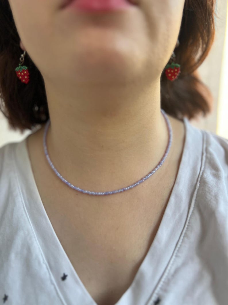 K&ouml;nnte beinhalten: Eine zarte, hellviolette Perlenkette wird getragen. Die Kette wird mit Erdbeerohrringen kombiniert. Die Ohrringe sind rot mit gr&uuml;nen Bl&auml;ttern und wei&szlig;en Punkten. Die Person tr&auml;gt ein wei&szlig;es Hemd.