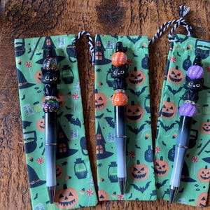 May include: Three pens with black-to-grey gradient barrels, each topped with a black cat figure between two patterned beads. The pens are displayed on individual green fabric pouches featuring a repeating pattern of jack-o'-lanterns, bats, and hats.