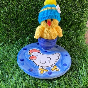 May include: A yellow knitted chick wearing a blue and yellow hat with a white flower, perched on a blue ceramic base. The base features a hand-painted white chicken with red accents and blue polka dots.
