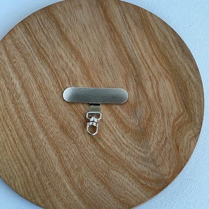 Könnte beinhalten: Ein silberfarbenes Metallbeschlagteil mit einem flachen, länglichen rechteckigen Riegel, der an einem kleinen, drehbaren Karabinerverschluss befestigt ist. Die Komponente hat eine glatte, reflektierende Oberfläche und dient zur Befestigung an anderen Gegenständen.
