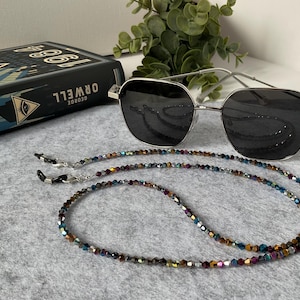 Könnte beinhalten: Eine silberfarbene Sonnenbrille mit dunklen Gläsern und einer langen Perlenkette. Die Kette besteht aus kleinen, facettierten, mehrfarbigen Perlen in Blau-, Violett-, Gold- und Grüntönen. Die Sonnenbrille liegt neben einem Buch mit dem Titel 1984.