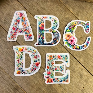 May include: Five colourful floral alphabet stickers with a white background. The letters are A, B, C, D, and E. Each letter is decorated with a different floral pattern.