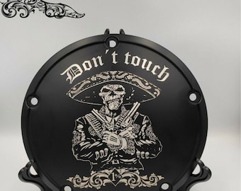 May include: Black motorcycle part featuring a skull in a sombrero holding two guns. The text "Don't touch" is above the skull. The design includes the text "1%" and decorative flourishes.