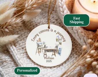 May include: A circular, white ceramic Christmas ornament with a gold hanging loop. The ornament displays a watercolour illustration of an artist's studio and the text "Every canvas is a journey." It is personalised with a name and the year 2025.