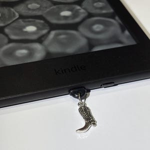 May include: A black Kindle e-reader with a silver cowboy boot charm attached. The screen shows a grayscale image. The word "kindle" is printed on the device.