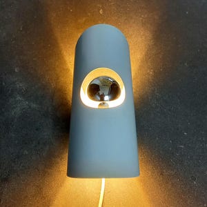 Könnte beinhalten: Hellblaue, zylindrische Wandleuchte mit abgerundeter Oberseite und kreisförmiger Leuchte. Die Leuchte hat eine reflektierende, verspiegelte Oberfläche. Ein weißes Kabel erstreckt sich von unten.