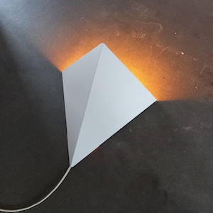 May include: A white pyramid-shaped lamp with a cord hanging down. The lamp is casting a warm yellow light on a dark surface.