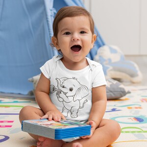 May include: A baby wearing a white onesie with a black and white illustration of an elephant playing a trumpet. The baby is sitting on a colorful play mat and holding a book.