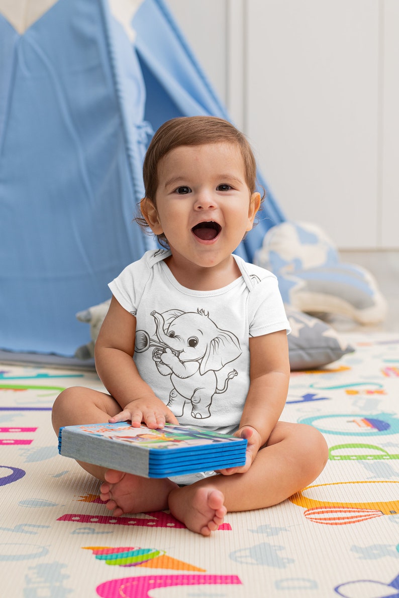 May include: A baby wearing a white onesie with a black and white illustration of an elephant playing a trumpet. The baby is sitting on a colorful play mat and holding a book.