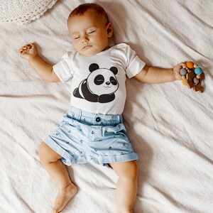 Pu&ograve; includere: Un bambino che indossa un body bianco a maniche corte con una stampa di panda bianco e nero. Il body ha il testo "G" in basso. Il bambino indossa pantaloncini blu con un motivo di ancora bianca. Il bambino &egrave; sdraiato su una coperta bianca e tiene un giocattolo di legno con perline colorate.