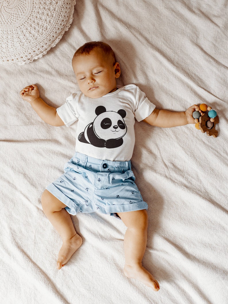Pu&ograve; includere: Un bambino che indossa un body bianco a maniche corte con una stampa di panda bianco e nero. Il body ha il testo "G" in basso. Il bambino indossa pantaloncini blu con un motivo di ancora bianca. Il bambino &egrave; sdraiato su una coperta bianca e tiene un giocattolo di legno con perline colorate.
