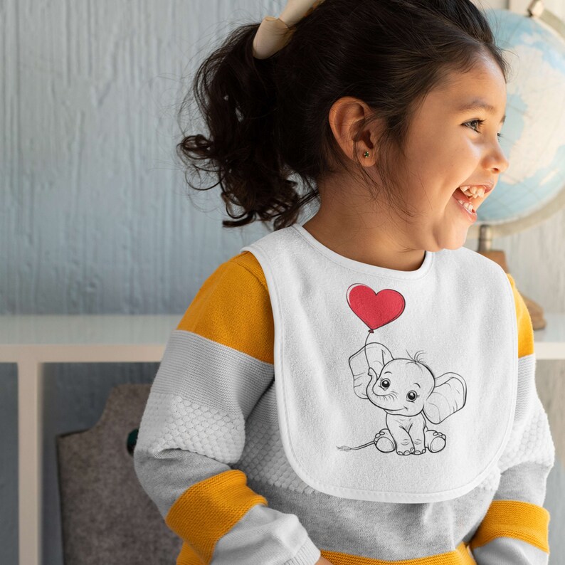 May include: A white bib featuring a black and white illustration of an elephant holding a red heart-shaped balloon. The bib is being worn by a young child with dark hair.
