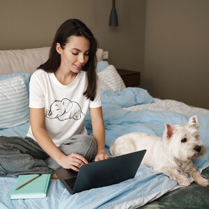 May include: A woman sits on a bed with a white dog and a laptop. She is wearing a white t-shirt with a black and white illustration of a sleeping elephant. The dog is white with a black nose and black eyes. The bed is covered in a blue blanket.