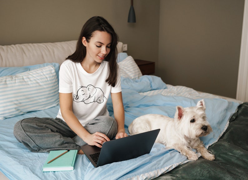 May include: A woman sits on a bed with a white dog and a laptop. She is wearing a white t-shirt with a black and white illustration of a sleeping elephant. The dog is white with a black nose and black eyes. The bed is covered in a blue blanket.