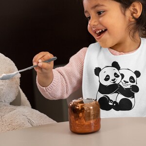 Pu&ograve; includere: Un bavaglino bianco per neonati con un'illustrazione in bianco e nero di due panda che si abbracciano. Un bambino piccolo indossa il bavaglino e sta nutrendo un giocattolo di peluche con un cucchiaio.