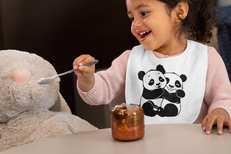 Pu&ograve; includere: Un bavaglino bianco per neonati con un'illustrazione in bianco e nero di due panda che si abbracciano. Un bambino piccolo indossa il bavaglino e sta nutrendo un giocattolo di peluche con un cucchiaio.