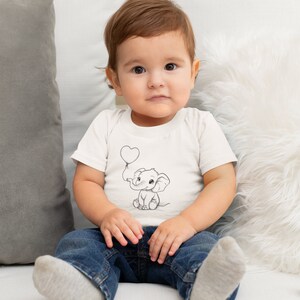May include: A young child wearing a white t-shirt with a black and white illustration of an elephant holding a heart-shaped balloon. The child is sitting on a white couch wearing blue jeans and white socks.