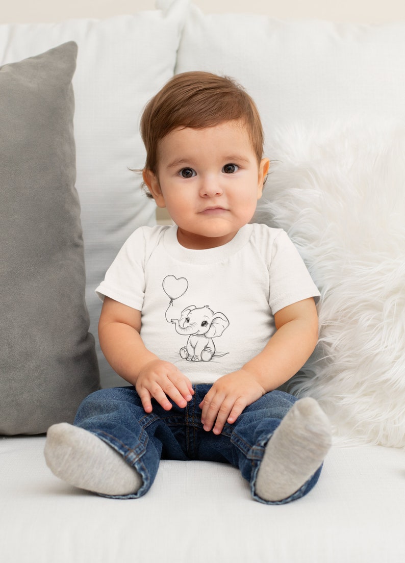 May include: A young child wearing a white t-shirt with a black and white illustration of an elephant holding a heart-shaped balloon. The child is sitting on a white couch wearing blue jeans and white socks.