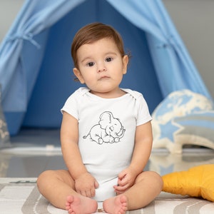 May include: A baby wearing a white onesie with a black and white illustration of a sleeping elephant. The baby is sitting on a gray and white patterned rug in front of a blue play tent.