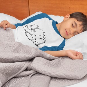 May include: A young person wearing a white and blue shirt with a black and white illustration of an elephant sleeping on the front. The person is sleeping on a white pillow and a gray blanket.