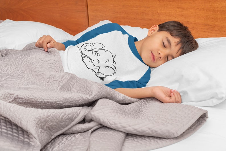 May include: A young person wearing a white and blue shirt with a black and white illustration of an elephant sleeping on the front. The person is sleeping on a white pillow and a gray blanket.