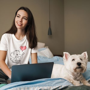 May include: A woman sitting on a bed with a laptop and a white dog. She is wearing a white t-shirt with a cartoon elephant holding a heart-shaped balloon. The dog is looking at the camera.
