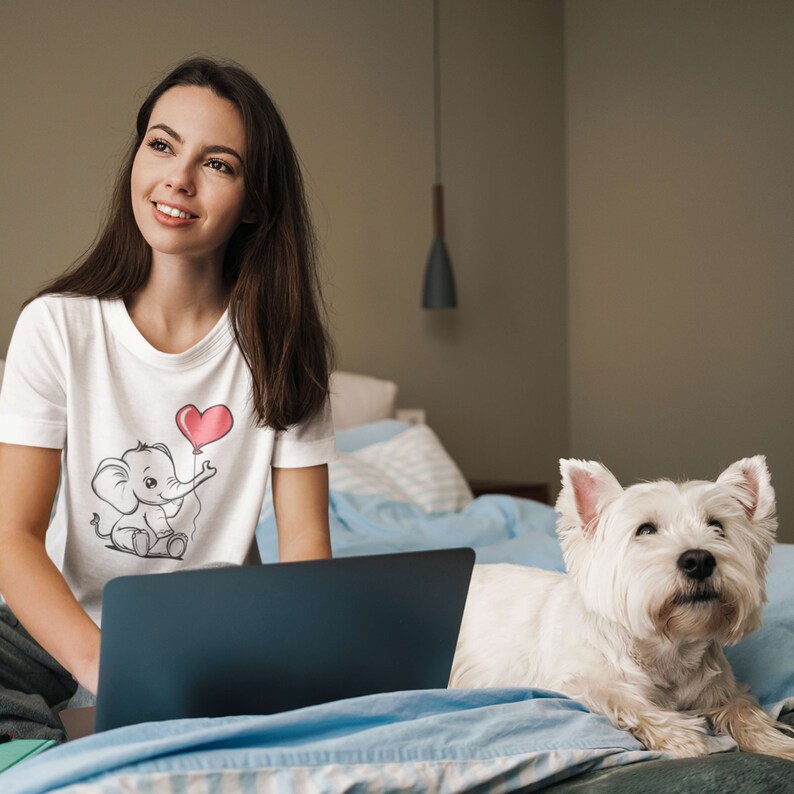 May include: A woman sitting on a bed with a laptop and a white dog. She is wearing a white t-shirt with a cartoon elephant holding a heart-shaped balloon. The dog is looking at the camera.