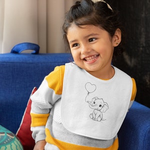 May include: A young child wearing a white bib with a black and white illustration of an elephant holding a heart-shaped balloon. The bib has a yellow and white striped shirt underneath.