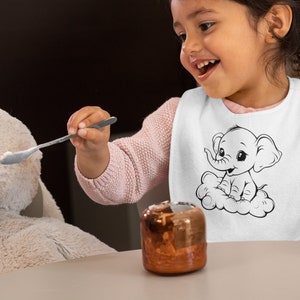 May include: A young child wearing a white bib with a black and white illustration of an elephant sitting on a cloud. The bib has the text "Elephant" on it. The child is holding a spoon and feeding a plush toy.
