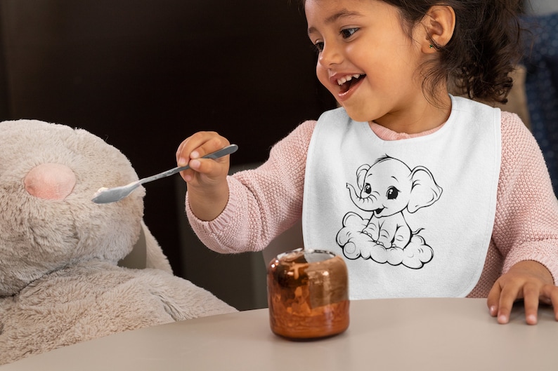 May include: A young child wearing a white bib with a black and white illustration of an elephant sitting on a cloud. The bib has the text "Elephant" on it. The child is holding a spoon and feeding a plush toy.