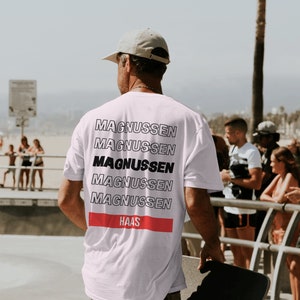 Puede incluir: Camiseta blanca con el nombre "MAGNUSSEN" apilado en un dise&ntilde;o repetitivo en la espalda. La palabra "MAGNUSSEN" est&aacute; en negrita negra. Una franja horizontal roja con la palabra "HAAS" est&aacute; debajo. La persona sostiene una patineta.