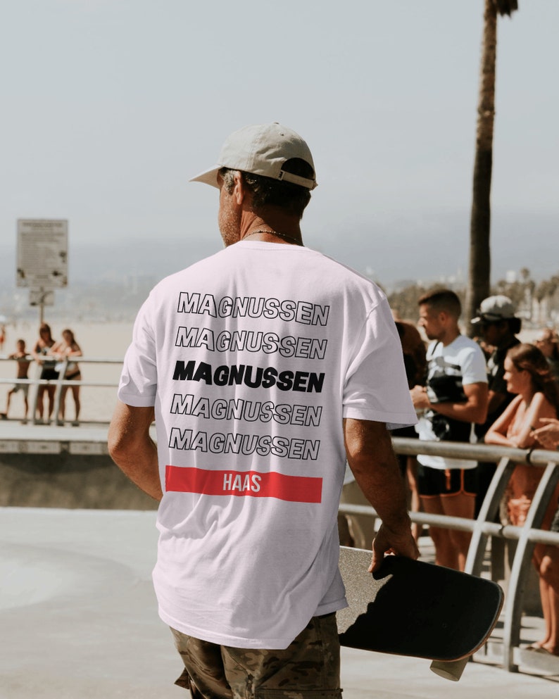 Puede incluir: Camiseta blanca con el nombre "MAGNUSSEN" apilado en un dise&ntilde;o repetitivo en la espalda. La palabra "MAGNUSSEN" est&aacute; en negrita negra. Una franja horizontal roja con la palabra "HAAS" est&aacute; debajo. La persona sostiene una patineta.