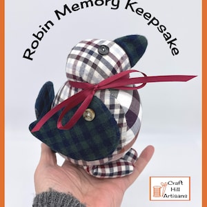 May include: A handmade plaid fabric robin keepsake with a red ribbon and button eyes. The robin is sitting in a person's hand. The text "Robin Memory Keepsake" is written in a circle around the robin. The text "Craft Hill Artisans" is in the bottom right corner of the image.