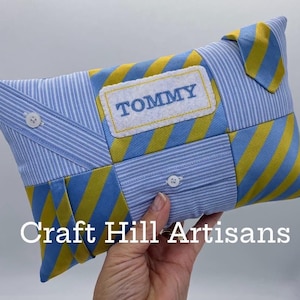 May include: A patchwork cushion made from blue and white striped fabric and yellow and blue striped fabric. The cushion has a white patch embroidered with the name "TOMMY" in blue. The cushion is held by a hand.