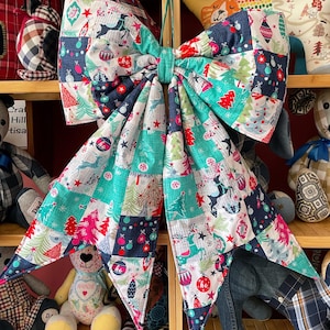 May include: A large decorative bow made of fabric with a patchwork pattern featuring Christmas trees, reindeer, ornaments, and the text ho ho. The fabric has a colour palette of teal, navy blue, red, and white, with two long hanging tails.