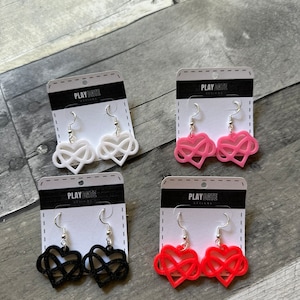 May include: Four pairs of heart-shaped earrings in white, pink, black, and red. Each earring features an infinity symbol within a heart shape, hanging from a silver hook. The earrings are displayed on white cards with the text "PLAY DATE DESIGNS".