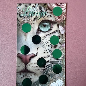 Könnte beinhalten: Eine rechteckige Karte mit einer detaillierten Illustration eines weißen Leoparden mit auffälligen blauen Augen, umgeben von floralen Akzenten und Schmetterlingen. Die Karte ist mit neun runden grünen Folienaufklebern und zwei weißen Würfeln versehen.