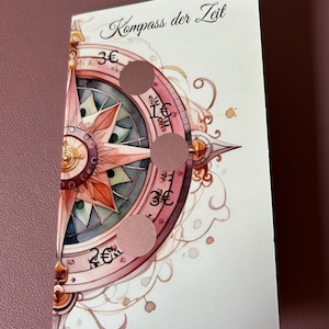 Könnte beinhalten: Eine weiße Karte mit einem rosafarbenen Kompass-Design. Der Kompass hat eine rosafarbene und blaue Mitte mit einem goldenen Pfeil. Die Karte trägt den Text "Kompass der Zeit" oben und hat vier Kreise mit Euro-Symbolen und Zahlen.