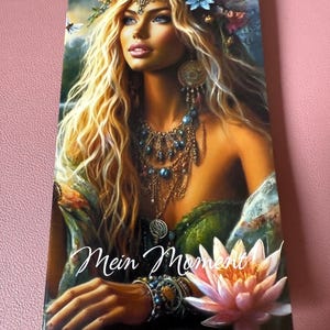 Könnte beinhalten: Porträt einer Frau mit langen blonden Haaren, geschmückt mit einem Blumenkranz und Schmuck. Das Bild zeigt den Text "Mein Moment" und eine rosa Lotusblume. Die Frau hat blaue Augen und trägt ein grünes Oberteil.
