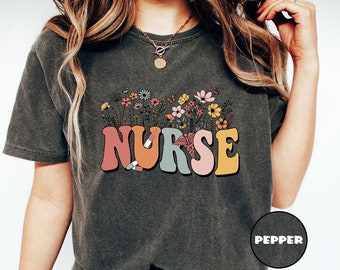 May include: A dark gray t-shirt with a retro-style graphic of the word "NURSE" in colorful letters surrounded by flowers and butterflies. The shirt is made of a soft, comfortable material and has a relaxed fit.