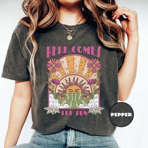Puede incluir: Una camiseta gris oscuro con un gráfico de estilo vintage de un sol saliendo detrás de flores y nubes. El texto "Here Comes The Sun" está impreso en la camiseta.