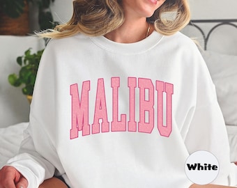 Op de afbeelding: Witte trui met het woord "MALIBU" bedrukt in roze letters. De trui wordt gedragen door een persoon die op een wit bed zit.
