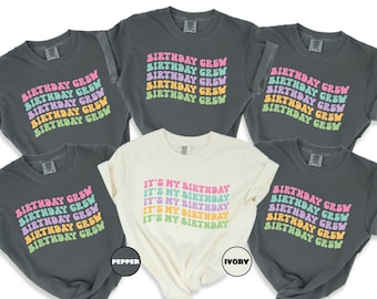 May include: Five gray t-shirts with the text "Birthday Crew" in rainbow colors. One white t-shirt with the text "It's My Birthday" in rainbow colors. The shirts are laid out on a white background.