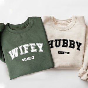 May include: Two sweatshirts, one green and one beige, with the words "WIFFY EST. 2024" and "HUBBY EST. 2024" printed on them in a varsity style font.