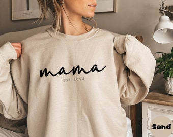 Könnte beinhalten: Ein beiger Pullover mit dem Text "mama est 2024" in schwarzer Schrift.
