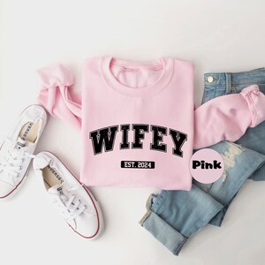 May include: Pink sweatshirt with the text "WIFEY EST. 2024" in black. The sweatshirt is laid out on a white surface with a pair of white sneakers and blue jeans.